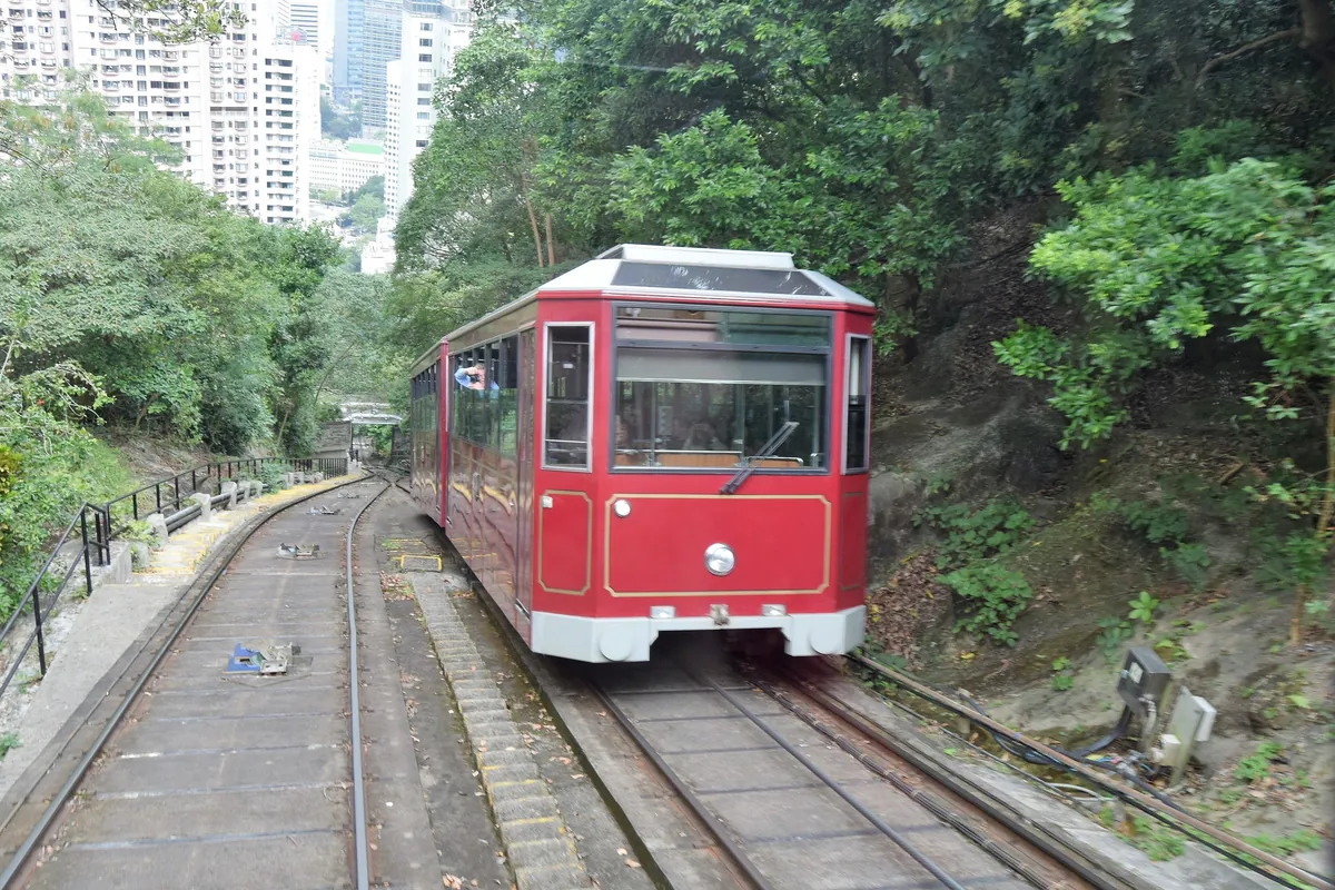 An earlier-generation red Peak Tram car approaching the passing loop on the wooded slope of Victoria Peak